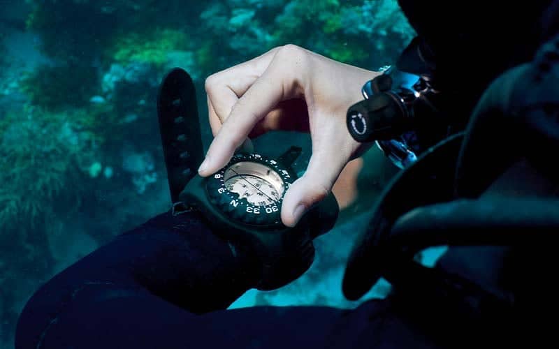 Diver checking a wrist-mounted dive compass underwater over a reef